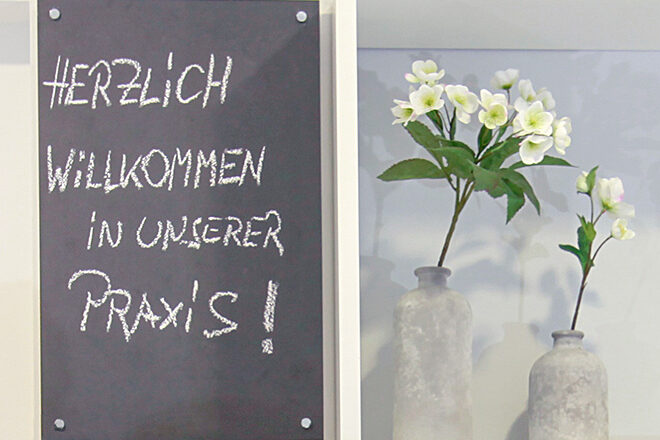 Eine Tafel mit der Aufschrift »Herzlich willkommen in unserer Praxis«, daneben eine Dekoration mit Blumen